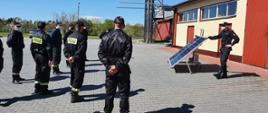Zdjęcie przedstawia strażaków stojących na placu KP PSP Opatów w trakcie szkolenia z instalacji fotowoltaicznych. W tle znajduje się szkoleniowy panel fotowoltaiczny oraz budynki komendy.