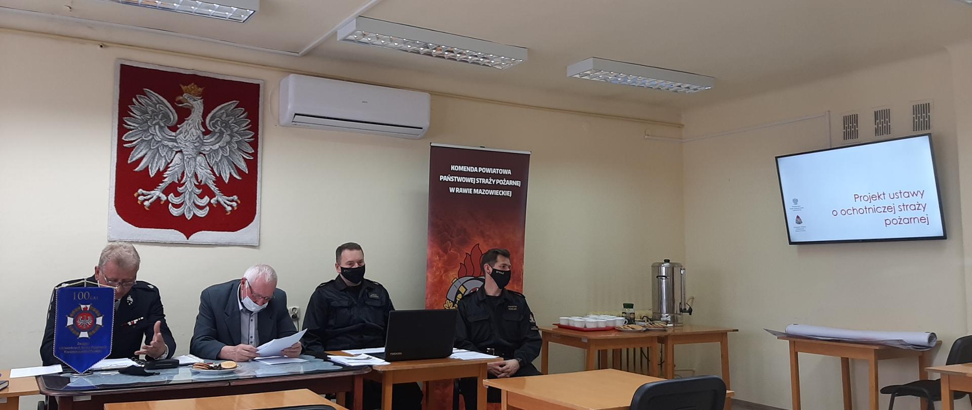 Zdjęcie przedstawia 4 mężczyzn siedzących za stołem, za nimi na ścianie wisi godło Polski oraz rozstawiony jest rolap Komendy Powiatowej Państwowej straży Pożarnej. Na stole przed nimi stoi laptop oraz dokumenty, które mężczyźni czytają. Na prawej ścianie znajduję się monitor z wyświetlaną prezentacją o Projekcie ustawy o ochotniczej straży pożarnej. Pierwszy mężczyzna od lewej ubrany jest w galowy strój osp, kolejny ubrany jest w garnitur z białą koszulą, natomiast kolejni dwaj mężczyźni ubrani są w stroje dowódczo-sztabowe. 