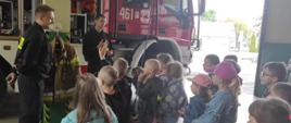 Wizyta uczniów w Komendzie Powiatowej PSP w Kozienicach. na pierwszym planie dzieci stojące tyłem do obiektywu, oglądające pokaz wozu strażackiego w garażu Komendy Powiatowej Państwowej Straży Pożarnej w Kozienicach , w tle strażcy przed samochodami pożarniczymi