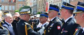 Strażak w mundurze galowym przypina odznaczenie strażakowi obok w rzędzie stoją inni strażacy oraz obok mężczyzna w garniturze i fotograf trzymający aparat.