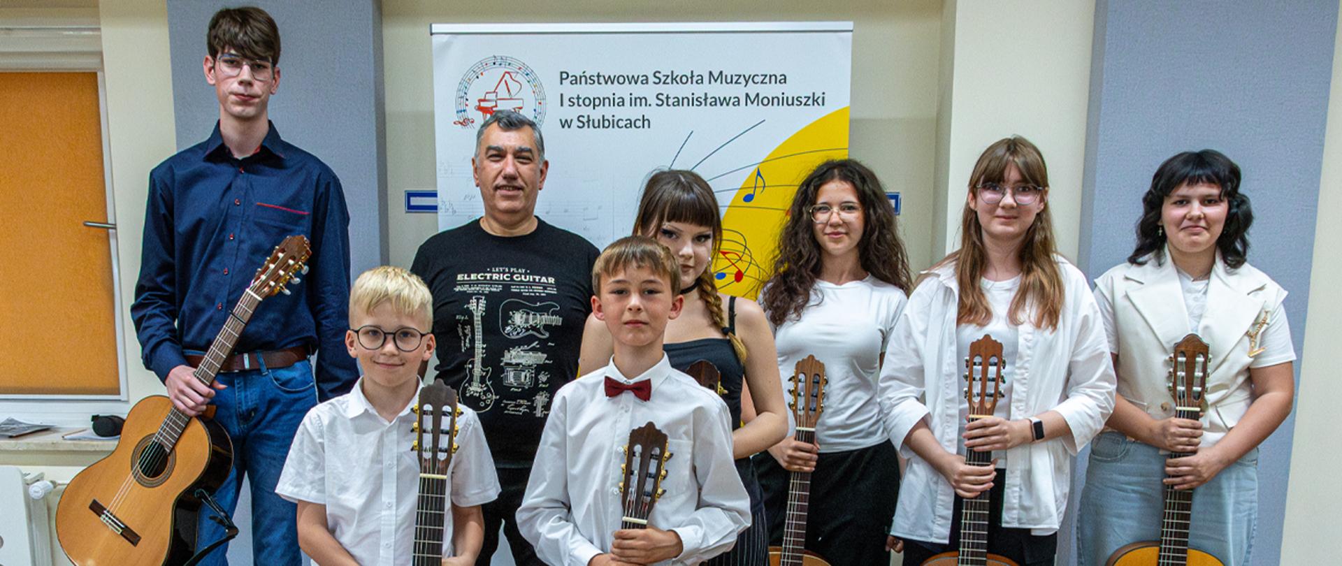 Na zdjęciu nauczyciel oraz uczniowie stoją w auli szkoły na tle szkolnego baneru. Uczniowie trzymają w rękach gitary.