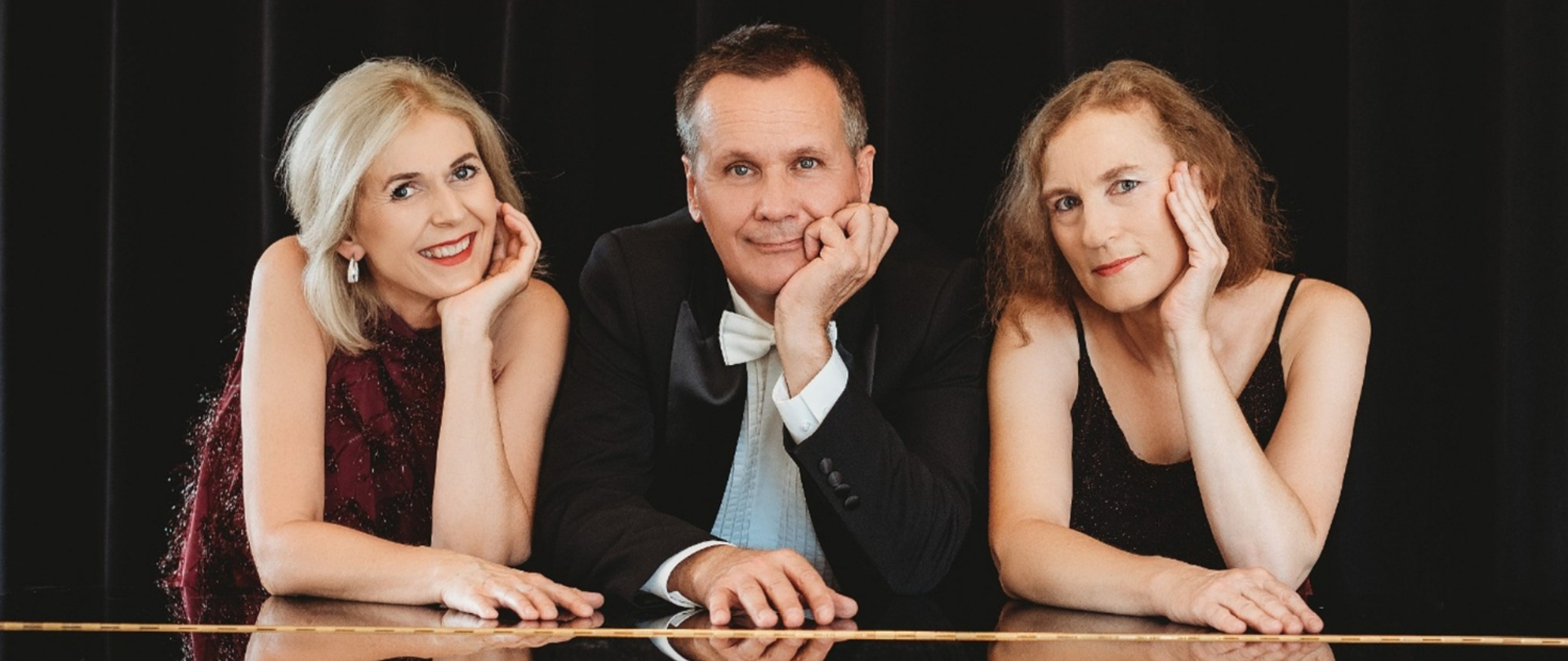 Plakat przedstawia informację nt koncertu Z miłości do muzyki. W centralnej części znajduje się zdjęcie trojga wykonawców opartych o fortepian, na górze tytuł koncertu, a na dole skład zespołu i program. Całośc jest na czerwonym tle. 