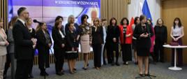Pod ścianą z pionowych drewnianych listewek z wbudowanym wielkim ekranem z napisem Powołanie Rady ds. Kobiet w Szkolnictwie Wyższym i Nauce stoi grupa kilkunastu kobiet, przed nimi stoją z dwóch stron sali minister Kulasek i wiceminister Zioło-Pużuk która mówi do mikrofonu na stojaku.