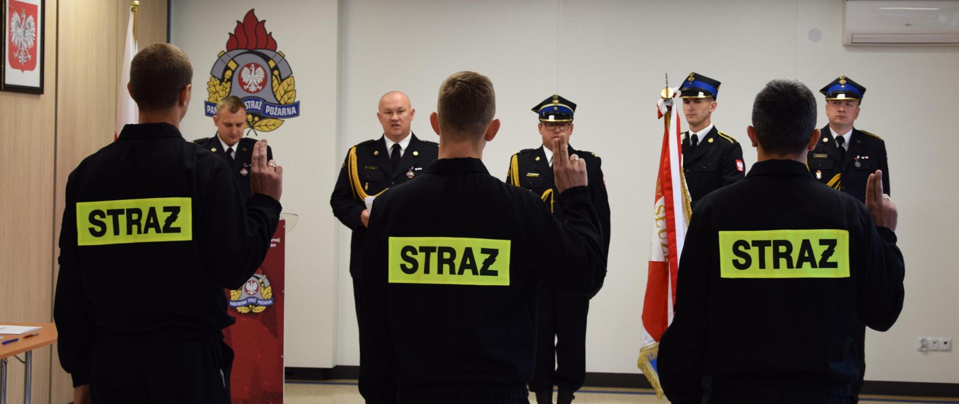 Nowo przyjęci funkcjonariusze ślubują na sztandar Komendy Powiatowej PSP w Mińsku Mazowieckim, Komendant Powiatowy PSP w Mińsku Mazowieckim odczytuje jego treść stojąc obok sztandaru, z prawej strony dowódca uroczystości, z lewej strony spiker.