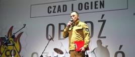  Funkcjonariusz straży pożarnej w piaskowym stroju – Komendant przemawia na scenie. W tle tło z logiem akcji „Czad i Ogień. Obudź Czujność”.