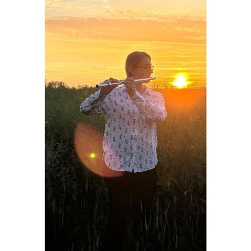 Zdjęcie kolorowe. Dziewczyna w białej koszuli i czarnych spodniach gra na flecie poprzecznym. W tle pole rzepaku i zachodzące słońce.