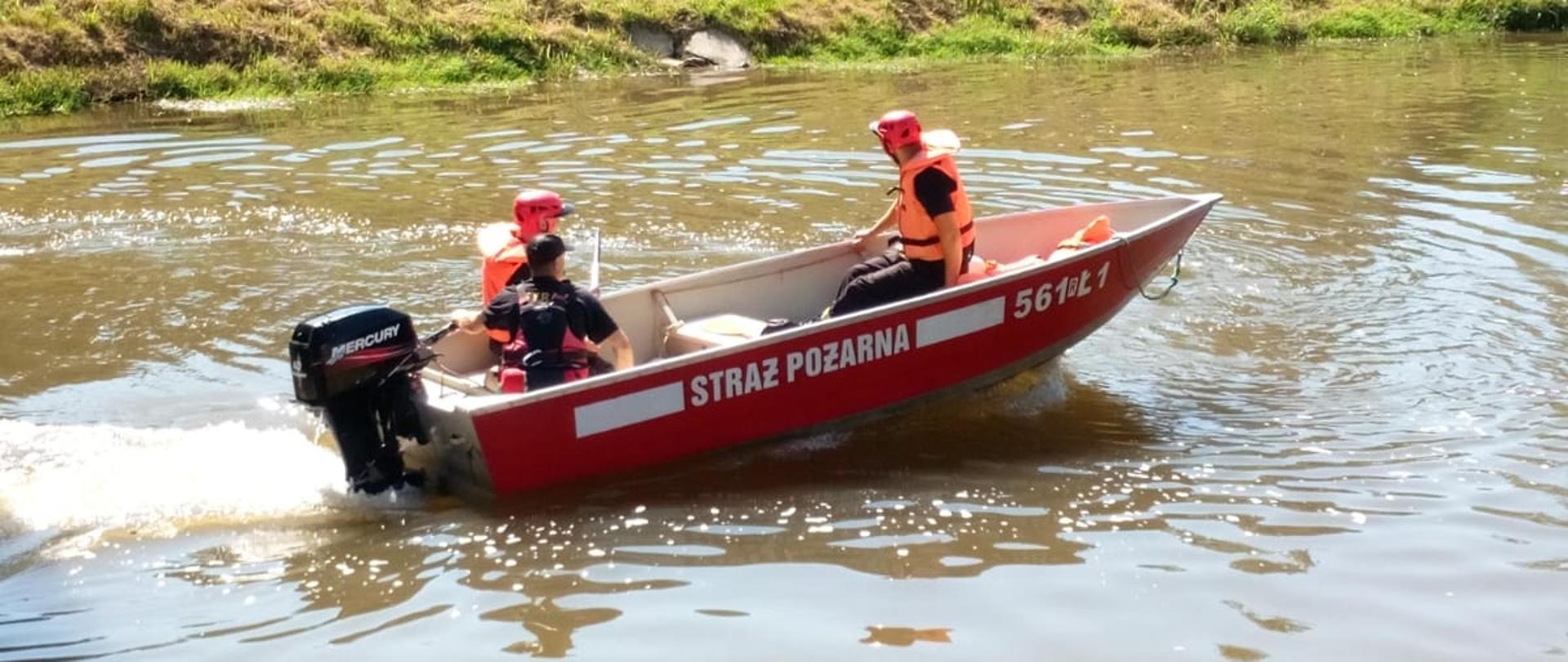 Na zdjęciu trzech funkcjonariuszy KP PSP w Strzyżowie płynie w łodzi po rzece Wisłok. Funkcjonariusze ubrani są w kamizelki asekuracyjne. W tle łąka oraz drzewa.