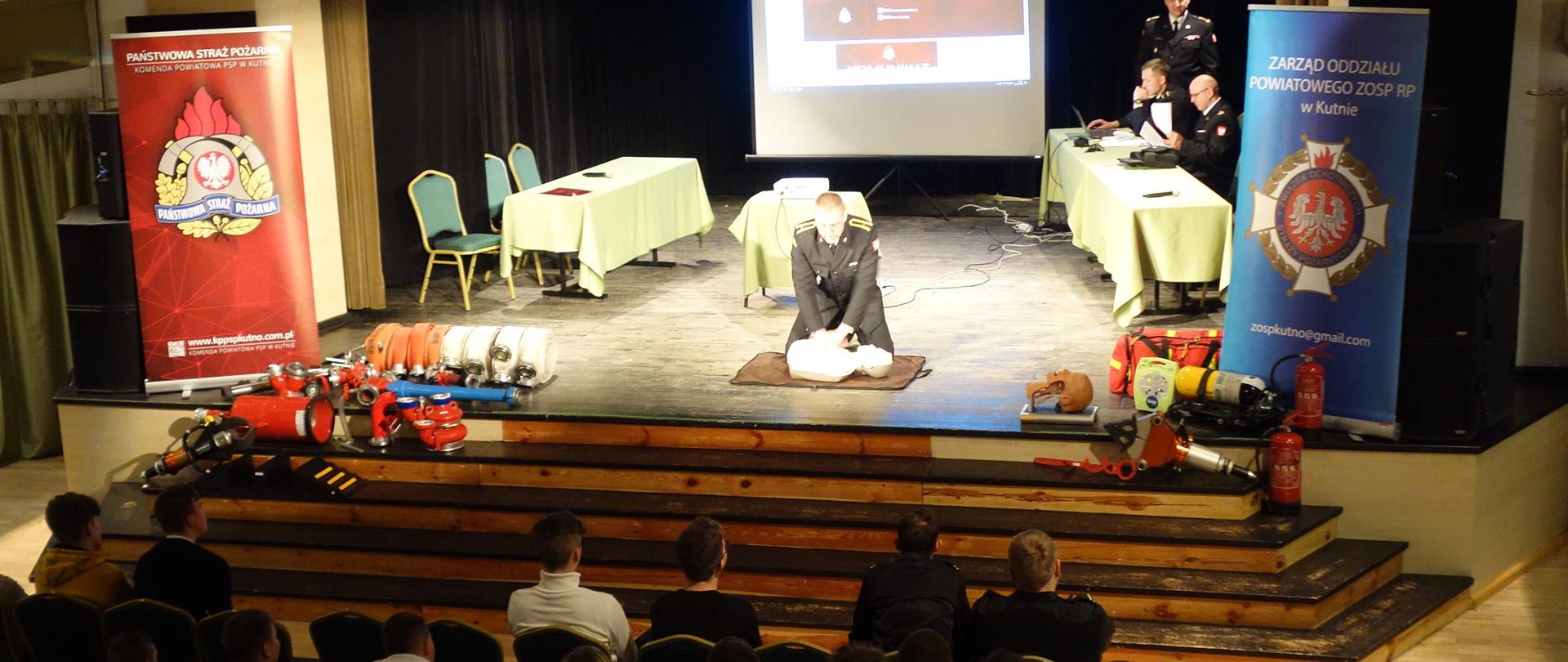 W sali odbywa się konkurs wiedzy pożarniczej. Na widowni siedzi młodzież. Na scenie strażak pokazujący na fantomie jak prawidłowo udzielać pierwszej pomocy.