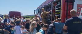 Na zdjęciu strażacy prezentują dzieciom wyposażenie samochodu ratowniczo-gaśniczego w ramach edukacyjnego spotkania plenerowego. Grupa młodych uczestników z zainteresowaniem słucha i przygląda się sprzętowi ratowniczemu. W tle widać również czerwony pojazd transportowy straży oraz malowniczy krajobraz letni.