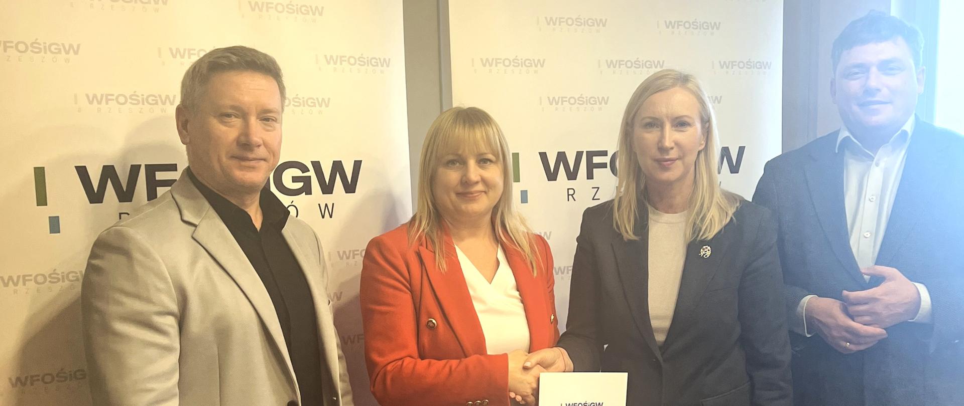 Na zdjęciu widać cztery osoby podczas oficjalnego spotkania w pomieszczeniu biurowym. Dwie kobiety siedzą przy drewnianym stole, a dwóch mężczyzn stoi za nimi. Tło stanowią dwa roll-upy z powtarzającym się logo „WFOŚiGW Rzeszów”. Po prawej stronie znajduje się okno, przez które wpada dzienne światło.
Na pierwszym planie, po lewej stronie stołu, siedzi kobieta w czerwonej marynarce i jasnej bluzce. Ma jasne, proste włosy z grzywką. Trzyma długopis i podpisuje dokument leżący na stole. Przed nią znajdują się kartki papieru oraz zielona pieczątka. Jej postawa jest wyprostowana, a wyraz twarzy spokojny i skupiony.
Po prawej stronie stołu siedzi druga kobieta ubrana w ciemną marynarkę i jasną bluzkę. Ma jasne włosy sięgające do ramion. Ręce ma złożone na dokumentach, które leżą przed nią. Na stole obok niej widoczny jest długopis oraz szklanka z wodą. Patrzy w stronę aparatu z delikatnym uśmiechem.
Za siedzącymi kobietami stoją dwaj mężczyźni w marynarkach. Mężczyzna po lewej ma jasną marynarkę i ciemną koszulę, stoi z rękami ułożonymi przed sobą i patrzy w obiektyw. Mężczyzna po prawej ma ciemną marynarkę i jasną koszulę; również patrzy w stronę aparatu, z lekko złożonymi dłońmi.
Na stole, oprócz dokumentów, widać także zamkniętą broszurę lub teczkę z nadrukiem godła oraz tekstem. Cała scena sugeruje formalne podpisanie umowy lub porozumienia w oficjalnym otoczeniu instytucjonalnym.