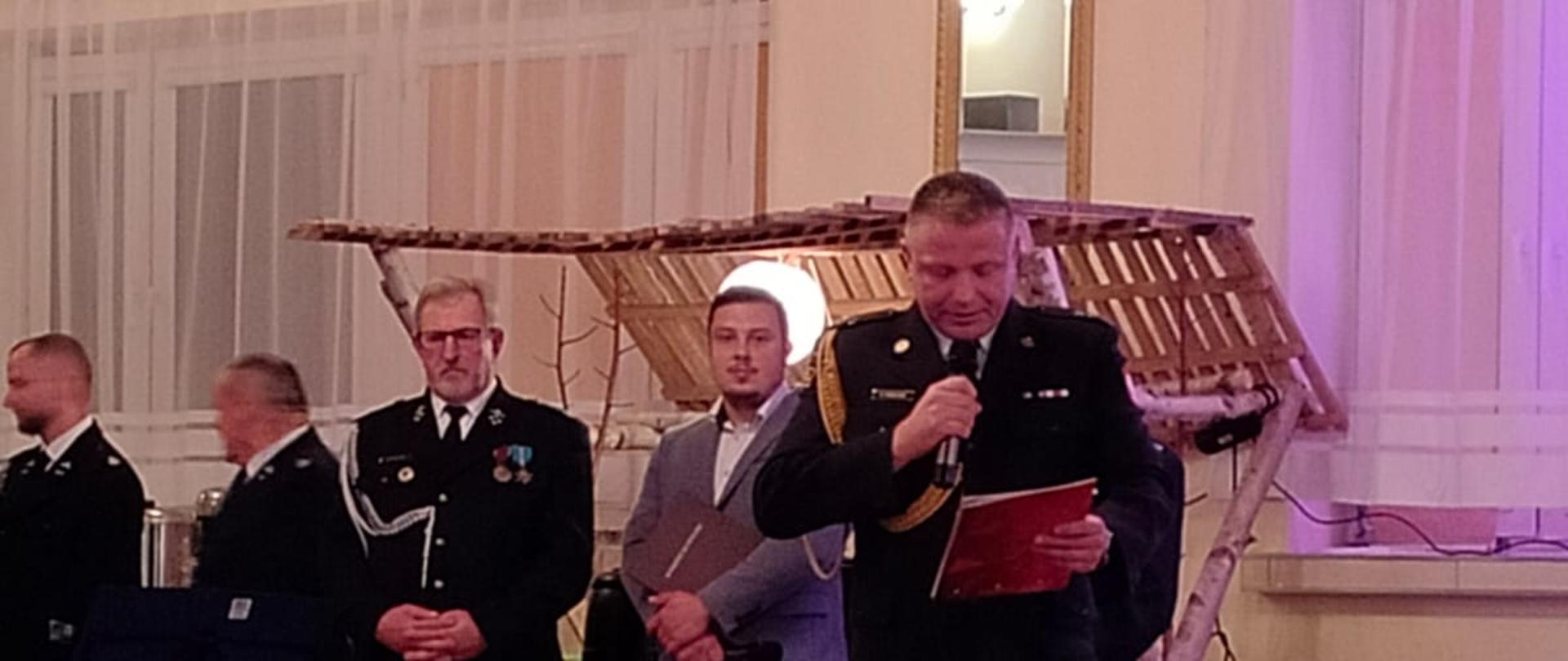 Zdjęcie wewnątrz Sali OSP Chełmce. Oświetlenie sztuczne. Widać członków orkiestry, OSP oraz funkcjonariuszy.. Drewniana podłoga oraz sufit.