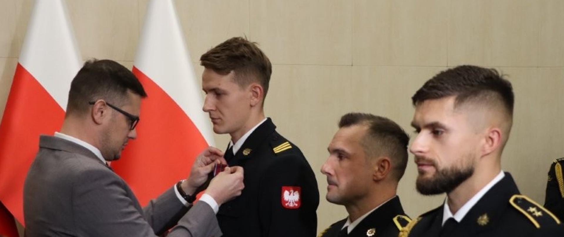 Z lewej Pan Wojewoda w siwym garniturze, z prawej trzej wyróżnieni strażacy w granatowych mundurach wyjściowych bez nakryć głowy. W tle dwie flagi Polski. Uroczystość przebiega w sali Urzędu. 