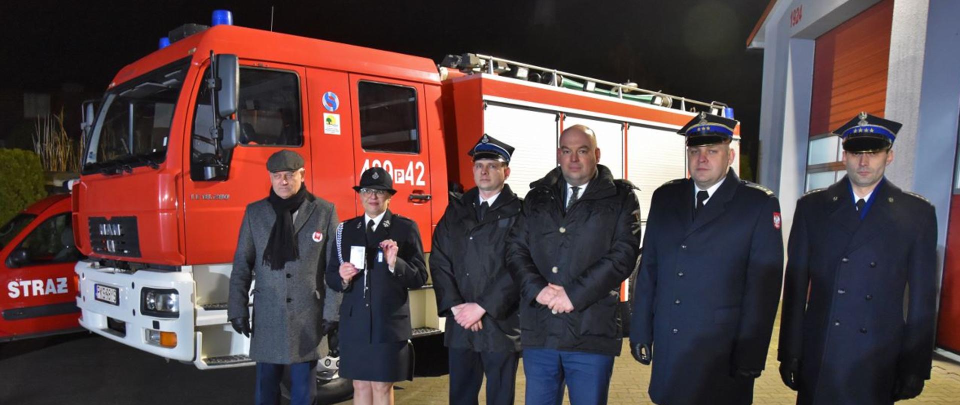 PRZEKAZANIE POJAZDÓW DLA JEDNOSTEK OSP. Na zdjęciu widać osoby i funkcjonariuszy PSP podczas symbolicznego przekazania kluczyków do samochodu, w tle samochód pożarniczy i strażnica OSP