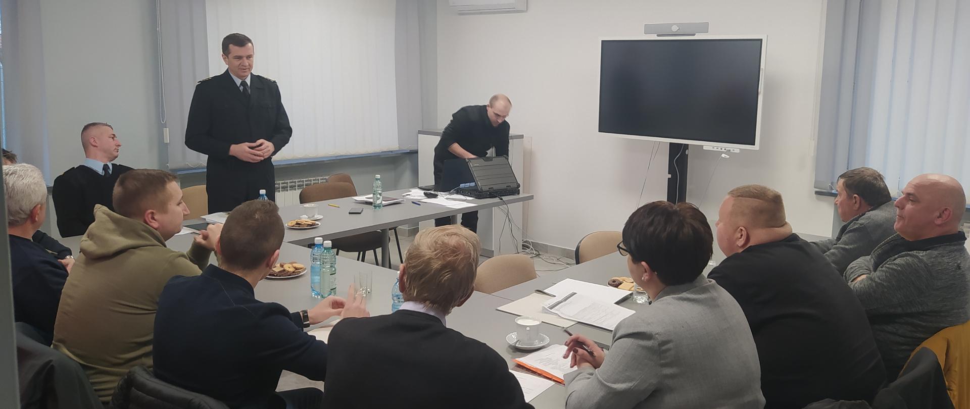 Zdjęcie przedstawia kilkanaście osób w pomieszczeniu konferencyjnym Komendy Powiatowej Państwowej Straży Pożarnej w Lesku. Jedna z osób - Komendant Powiatowy Państwowej Straży Pożarnej w Lesku, stojąc w umundurowaniu służbowym, przemawia do zebranych.