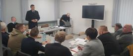 Zdjęcie przedstawia kilkanaście osób w pomieszczeniu konferencyjnym Komendy Powiatowej Państwowej Straży Pożarnej w Lesku. Jedna z osób - Komendant Powiatowy Państwowej Straży Pożarnej w Lesku, stojąc w umundurowaniu służbowym, przemawia do zebranych.