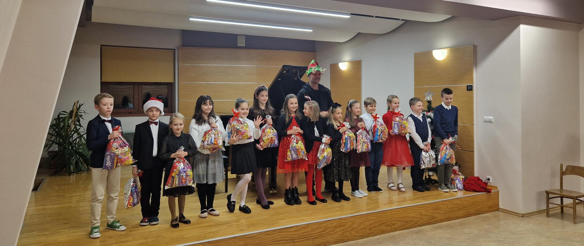 Uczniowie klasy drugiej sekcji instrumentów dętych i klawiszowych stoją w jednym rzędzie na scenie sali kameralnej. Dzieci odświętnie ubrane trzymają w dłoniach paczki mikołajkowe. Za dziećmi nauczyciel w w czapce mikołajowej. Za nimi fortepian, okno przysłonięte roletą. Pod oknem nawilżacz a obok nawilżacza z lewej strony palma doniczkowa. Z prawej strony na ścianie zapalone dwa kinkiety. Na suficie zapalone lampy. Z prawej strony puste krzesła.