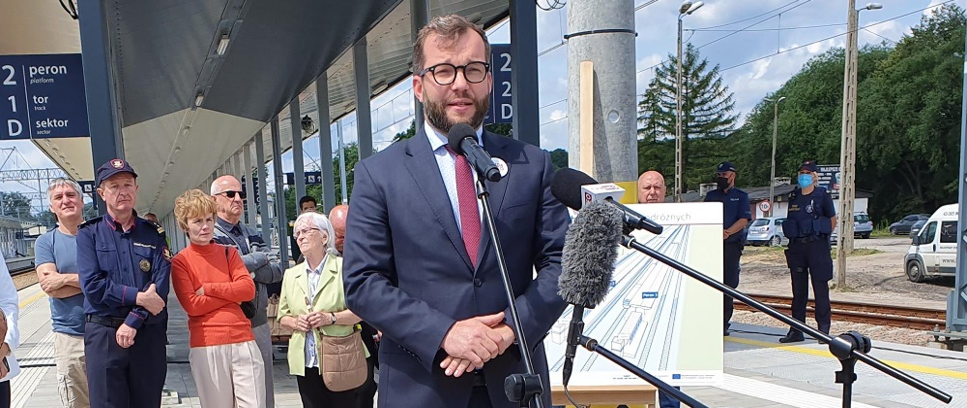 Minister Grzegorz Puda stoi przed mikrofonem podczas konferencji prasowej, w tle peron i grupa ludzie stojących