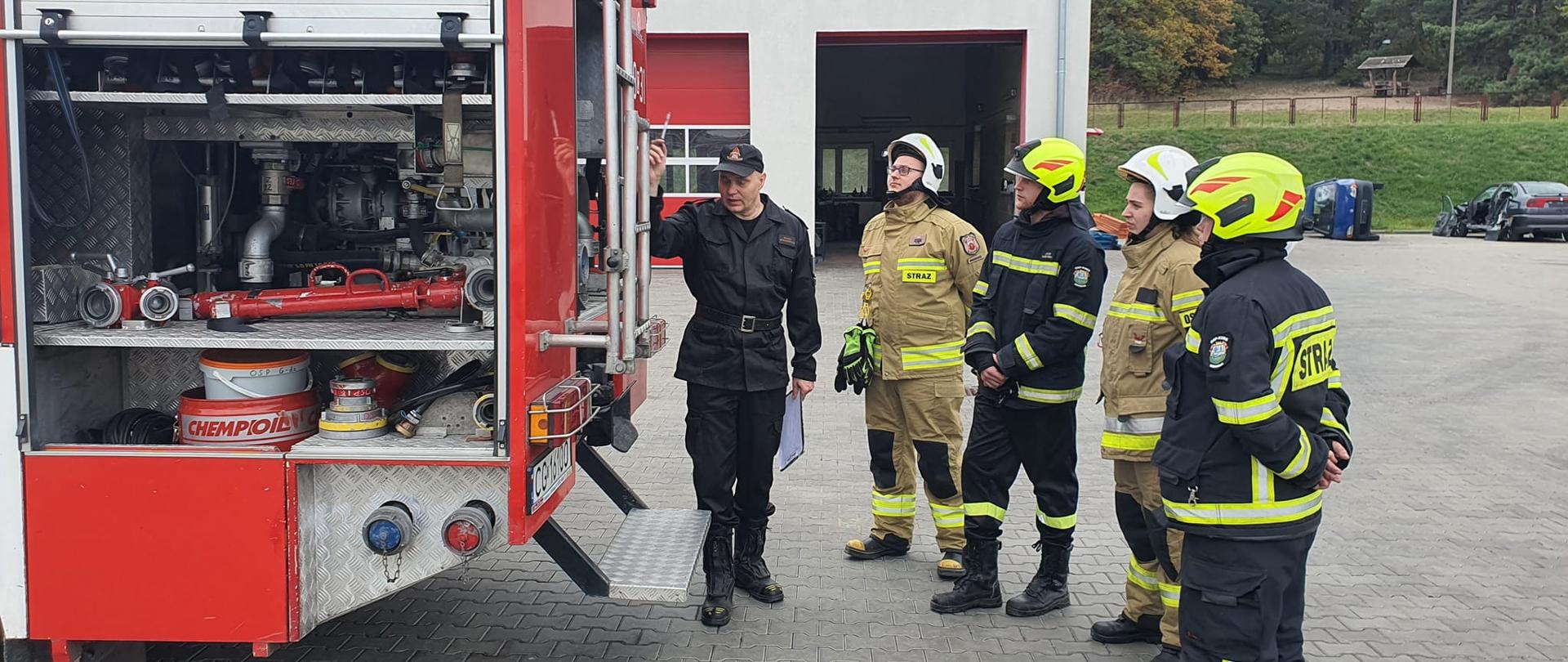 Zdjęcie przedstawia 4 strażaków w granatowych i piaskowych mundurach bojowych oraz białych i limonkowych hełmach, którzy stoją za tylną częścią czerwonego pojazdu bojowego. Strażak w czarnym mundurze przemawia do nich. W tle budynek z czerwonymi bramami oraz las.