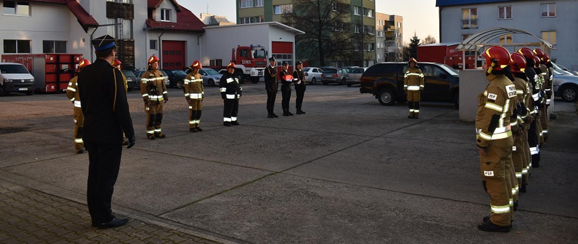 Uroczysta zmiana służby w JRG Zgorzelec. Funkcjonariusze ubrani w ubrania specjalne stoją na placu wewnętrznym KP w dwóch szeregach naprzeciw siebie. W głębi poczet falangowy w umundurowaniu galowym. Z lewej strony szyku stoi Z-ca Komendanta Powiatowego PSP w Zgorzelcu w umundurowaniu wyjściowym