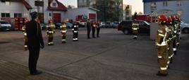 Uroczysta zmiana służby w JRG Zgorzelec. Funkcjonariusze ubrani w ubrania specjalne stoją na placu wewnętrznym KP w dwóch szeregach naprzeciw siebie. W głębi poczet falangowy w umundurowaniu galowym. Z lewej strony szyku stoi Z-ca Komendanta Powiatowego PSP w Zgorzelcu w umundurowaniu wyjściowym