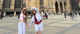 Panoramiczne zdjęcie dwóch osób stojących przed katedrą, unoszących ręce w kształt serca, z szerokim ujęciem placu i otoczenia.
