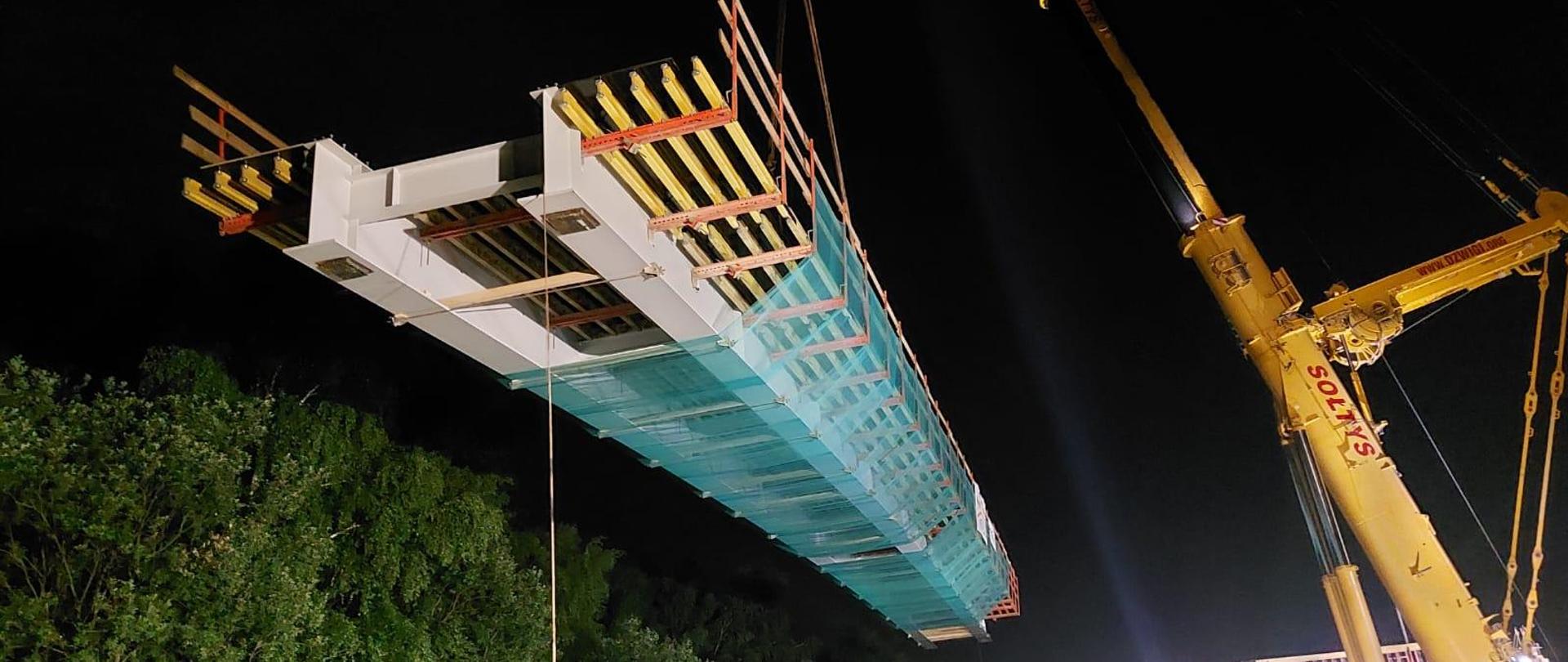 dźwig unosi przęsło kładki i ustawia ją nad podporami, prace nocne w Modlnicy