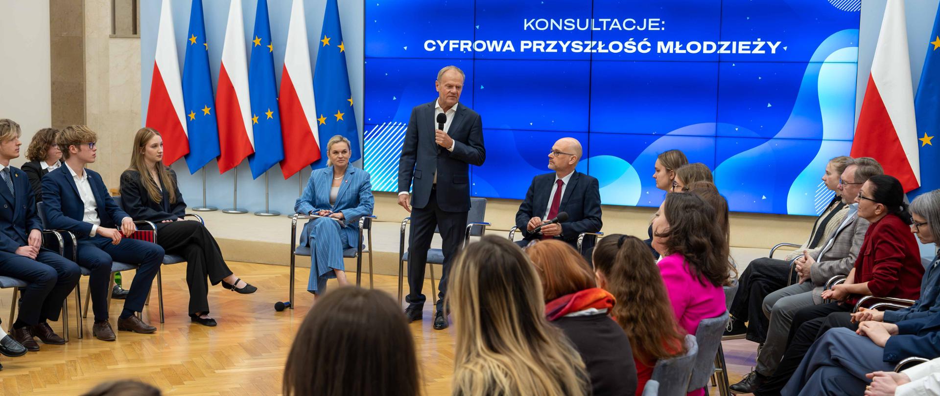 Szerokie ujęcie sali konferencyjnej podczas debaty. Premier Donald Tusk przemawia do zgromadzonej młodzieży siedzącej w kręgu. W tle widoczne są flagi Polski i Unii Europejskiej oraz duży ekran z hasłem konsultacji.