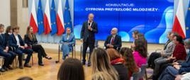 Szerokie ujęcie sali konferencyjnej podczas debaty. Premier Donald Tusk przemawia do zgromadzonej młodzieży siedzącej w kręgu. W tle widoczne są flagi Polski i Unii Europejskiej oraz duży ekran z hasłem konsultacji.