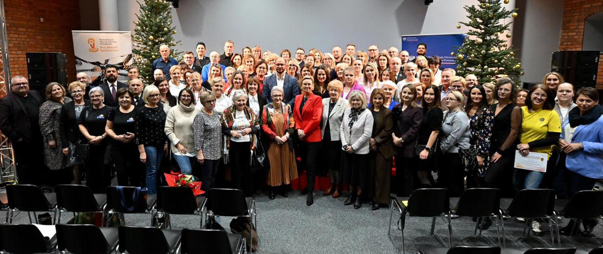 Szeroki kadr, zdjęcie grupowe. Uczestnicy konferencji pozują do wspólnego zdjęcia. W centralnej części zdjęcia widoczna Sekretarz Stanu Adriana Porowska oraz Michał Braun, dyrektor Narodowego Instytutu Wolności. W tle widoczne elementy wystroju sali: choinki przystrojone na święta, głośniki oraz banery promocyjne.