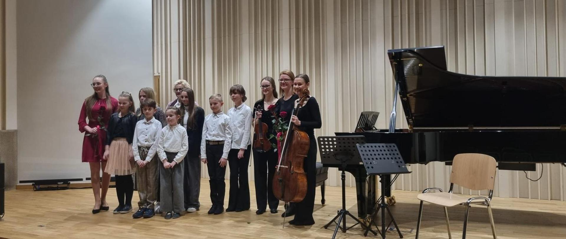 Na zdjęciu grupa osób odświętnie ubranych. Są to uczniowie, którzy wystąpili podczas koncertu. Obok nich stoi czarny fortepian. Przed fortepianem dwa czarne pulpity i brązowe krzesło. Tło obrazka w odcieniach brązu.