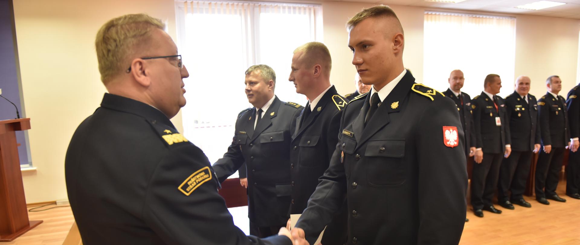 Na pierwszym planie Łódzki Komendant Wojewódzki gratuluje jednemu z młodszych aspirantów podjęcia stałej służby w szeregach Państwowej Straży Pożarnej. W tle stoją Komendanci Powiatowi Państwowej Straży Pożarnej.