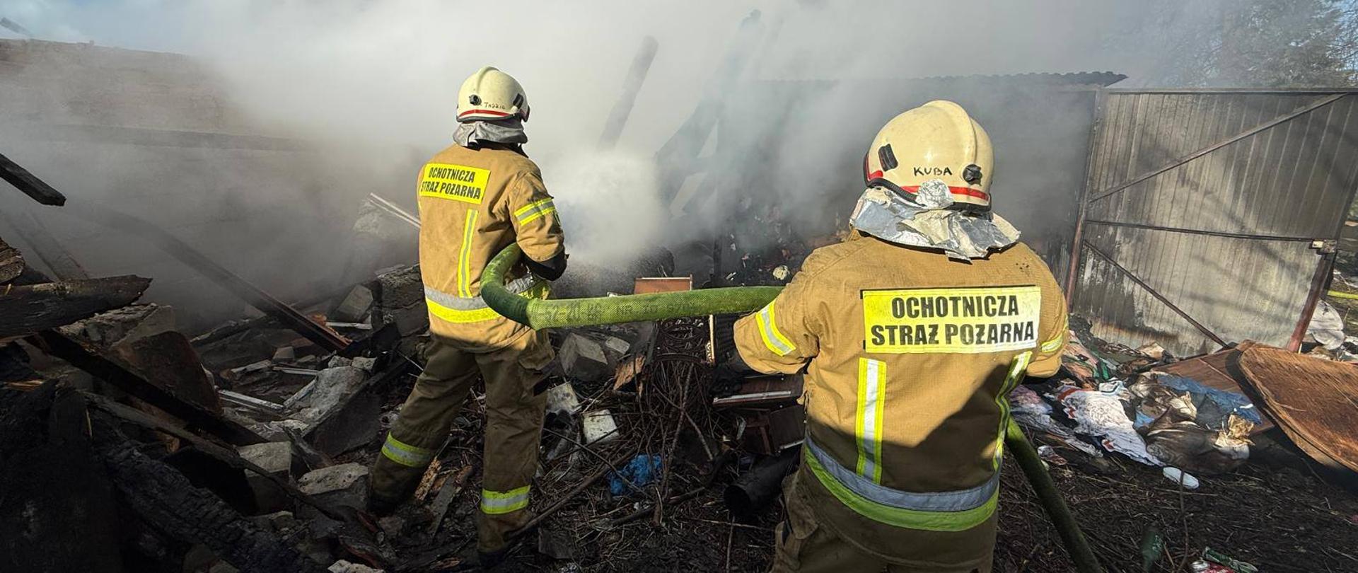 Dwóch strażaków stoi tyłem gaszą pożar, dużo dymu. Słonecznie.