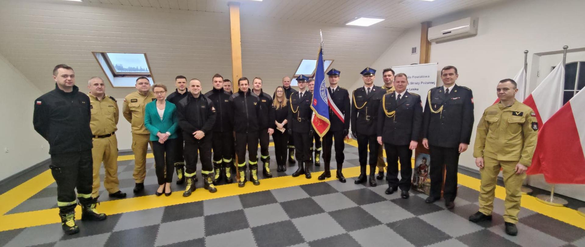 Zdjęcie ukazuje Strażaków i funkcjonariuszy cywilnych Komendy Powiatowej Państwowej Straży Pożarnej pozujących do pamiątkowego zdjęcia z odchodzącym funkcjonariuszem.
