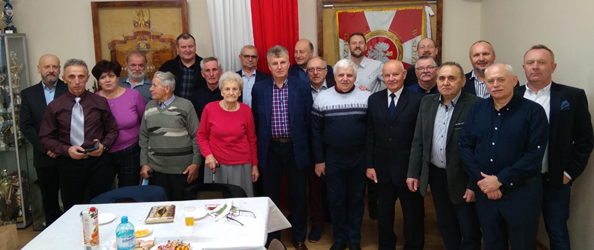 Zdjęcie grupowe członków koła związku emerytów i rencistów pożarnictwa. Na pierwszym planie zastanawiony stół z poczęstunkiem. Z grupą widoczny sztandar wiszący w gablocie oraz godło państwowe.