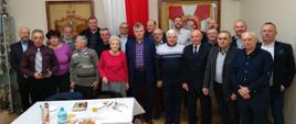 Zdjęcie grupowe członków koła związku emerytów i rencistów pożarnictwa. Na pierwszym planie zastanawiony stół z poczęstunkiem. Z grupą widoczny sztandar wiszący w gablocie oraz godło państwowe.