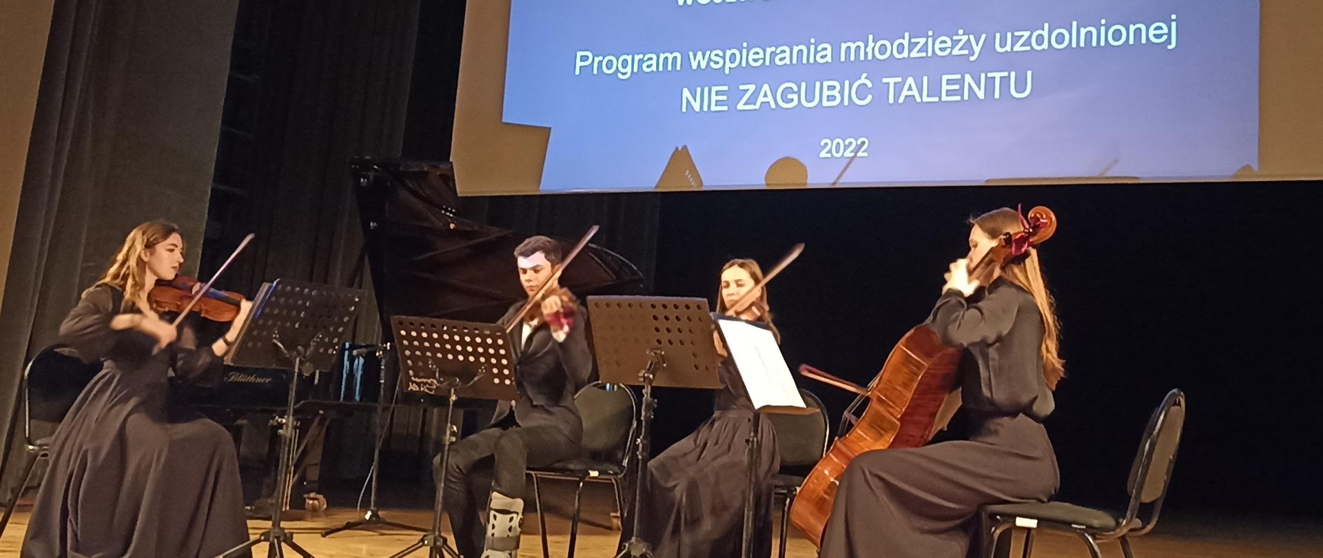 zdjęcie z gali wręczenie stypendiów w ramach programu wspierania młodzieży uzdolnionej „Nie zagubić talentu” przedstawiające kwartet smyczkowy PSM II stopnia pracujący pod opieką nauczyciela Ryszard Kuska.w składzie: Karolina Moskal - I skrzypce, Dawid Skóra - II skrzypce, Julia Chmiel - altówka, Hanna Kozioł - wiolonczela na scenie w Samorządowym Centrum Kultury w Mielcu.