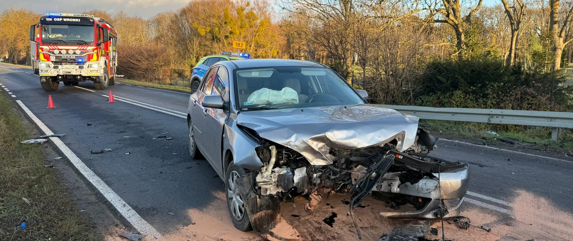 Uszkodzony srebrny samochód na drodze, za nim pojazd straży
