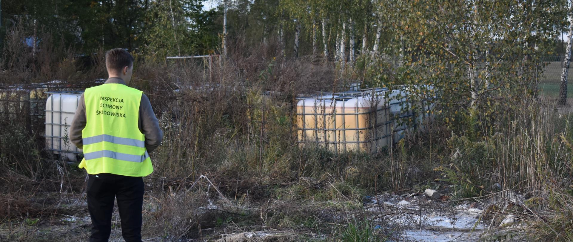 Inspektor Wojewódzkiego Inspektoratu Ochrony Środowiska w Warszawie stoi przed porzuconymi na zarośniętym terenie kilkoma zbiornikami o pojemności 1000 l.