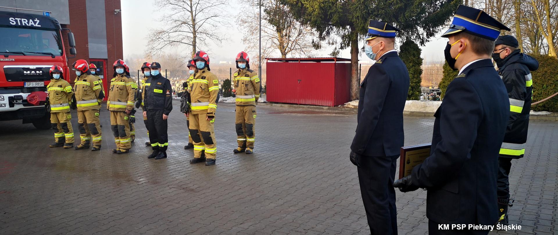 Zdjęcie przedstawia od lewej, fragment średniego samochodu ratowniczo - gaśniczego, 10 strażaków, ubranych w ubrania specjalne, stojących w dwuszeregu, komendanta miejskiego PSP w Piekarach Śląskich bryg. Tomasza Dziechciarza w mundurze wyjściowym z maseczką na twarzy, zastępcę komendanta miejskiego PSP w Piekarach Śląskich ml. bryg. Marcina Gąsowskiego oraz dowódcę JRG mł. bryg. Rafała Blachlińskiego. Wszyscy stoją na placu wewnętrznym Komendy.