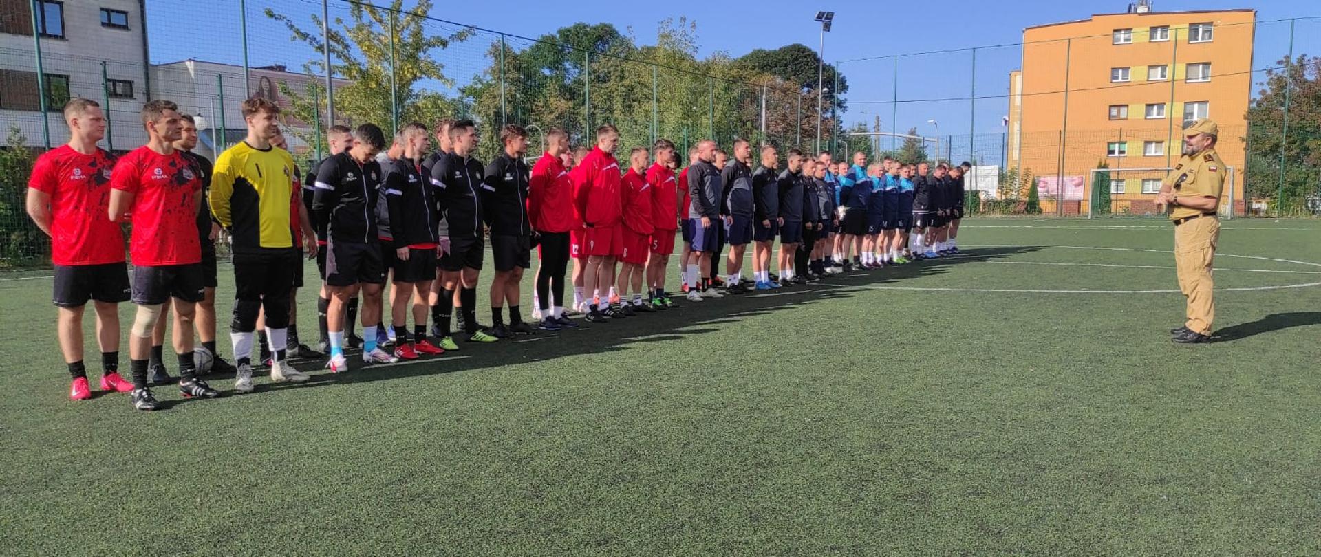 Na boisku grupa kilkudziesięciu zawodników w strojach piłkarskich. Przed nimi mężczyzna ubrany w ubranie służbowe strażackie.