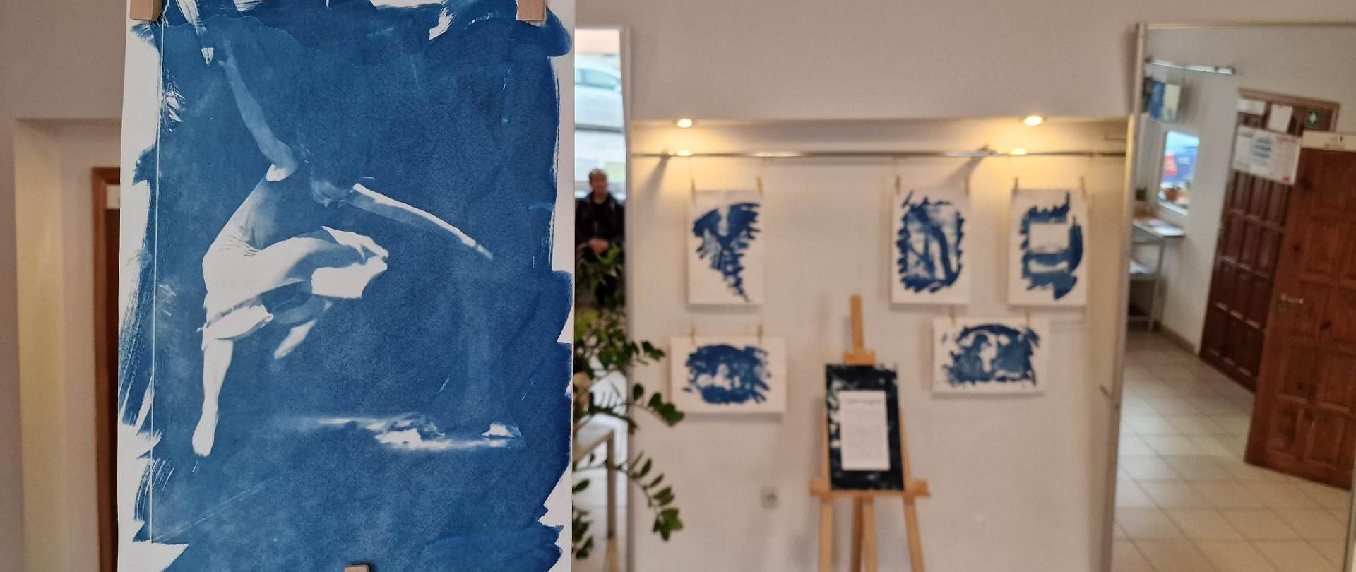 Zdjęcie prezentuje na pierwszym planie pracę artystyczną - zdjęcie wykonane w technice cyjanotypii. Praca jest w kolorze niebieskim, na pracy widnieje tancerka w pozie lotnej. W tle, rozmazana wystawa prac cyjanotypii. 