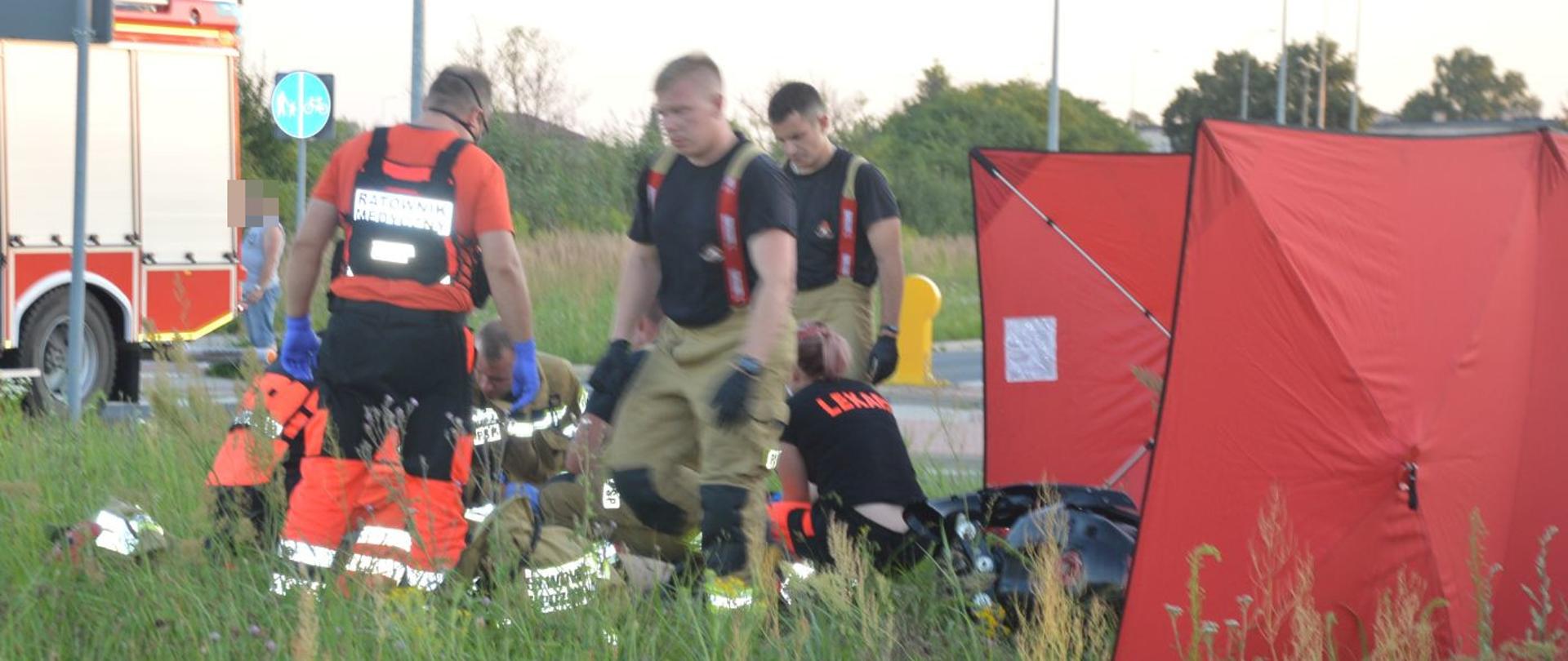 Śmiertelny wypadek motocyklisty. Rondo Bitew o Rawicz 1919. Na środku ronda strażacy i zespół ratownictwa medycznego ratujący życie młodego motocyklisty. Z prawej strony rozstawiony jest parawan ochronny, w tle wóz straży pożarnej.