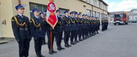 Fotografia z Dnia Strażaka w Komendzie Powiatowej Państwowej Straży Pożarnej w Mielcu. Zdjęcie wykonane podczas uroczystej akademii z okazji dnia strażaka w dniu 11 maja 2023 roku na placu jednostki ratowniczo-gaśniczej nr 2, komendy powiatowej Państwowej Straży Pożarnej w Mielcu. Na fotografii stoją funkcjonariusze ubrani w galowe mundury koloru granatowego. Jeden z strażaków trzyma Sztandar Komendy Powiatowej Państwowej Straży Pożarnej w Mielcu na którym widać godło państwowe.