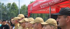 To zbliżenie przedstawia grupę strażaków Państwowej Straży Pożarnej (PSP) stojących w szeregu. Są to zarówno mężczyźni, jak i kobiety, ubrani w jasnobrązowe mundury z naszywkami i pagonami. Niektórzy noszą jasnobrązowe czapki z daszkiem. Osoba na pierwszym planie po prawej stronie, poza szeregiem, ma na sobie czarną czapkę z daszkiem i czarną koszulkę. W tle, za szeregiem strażaków, widać fragment czerwonego namiotu z napisem "KOMENDA POWIATOWA ... W OSTROWI MAZOWIECKIEJ", co wskazuje na miejsce wydarzenia. Po lewej stronie w tle widać również fragmenty zielonego lasu. Scena sugeruje apel lub uroczystą część zawodów.