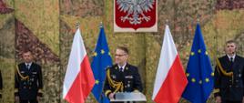 Komendant Morskiego Oddziału Straży Granicznej stoi przed mównicą i przemawia. Za nim stoi dwóch funkcjonariuszy Straży Granicznej, flagi Polski, Unii Europejskiej oraz wisi godło Polski.