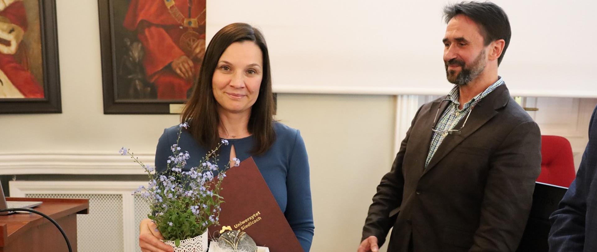 Kobieta pozuje do zdjęcia. Trzyma w dłoni doniczkę z kwiatami oraz medal (wyróżnienie). Obok niej stoi mężczyzna w garniturze. 