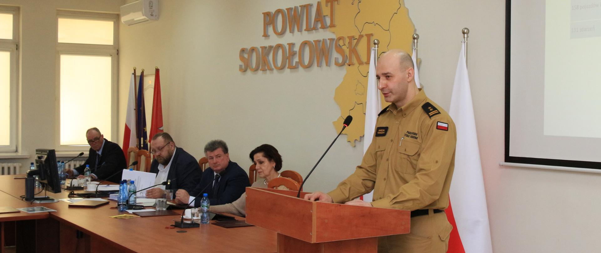 Złożenie informacji Komendanta Powiatowego PSP w Sokołowie Podlaskim dotyczącej bezpieczeństwa przeciwpożarowego powiatu sokołowskiego w 2023 roku - na zdjęciu komendant stoi za mównicą i przemawia w starostwie.