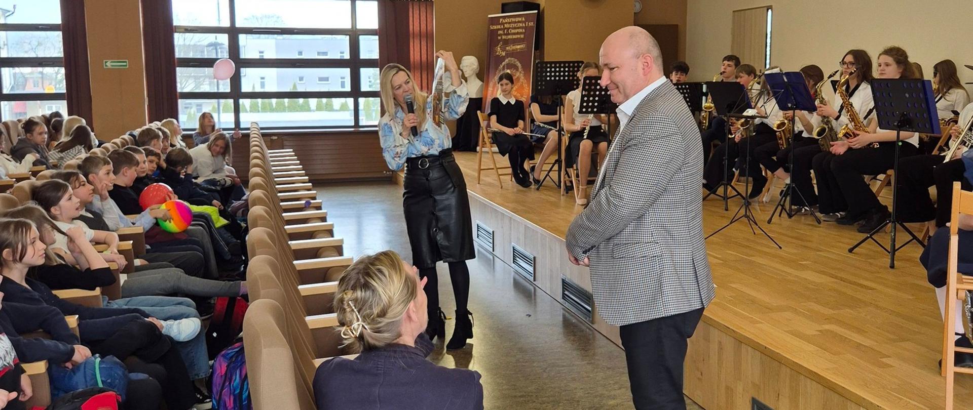 Tomasz Winczewski prowadzi koncert dla uczniów szkół podstawowych. W tle orkiestra i słuchacze.