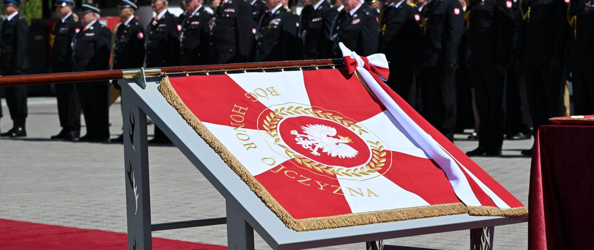 Biało czerwony sztandar leżący na stole. W tle strażacy podczas uroczystej zbiórki