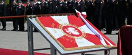 Biało czerwony sztandar leżący na stole. W tle strażacy podczas uroczystej zbiórki